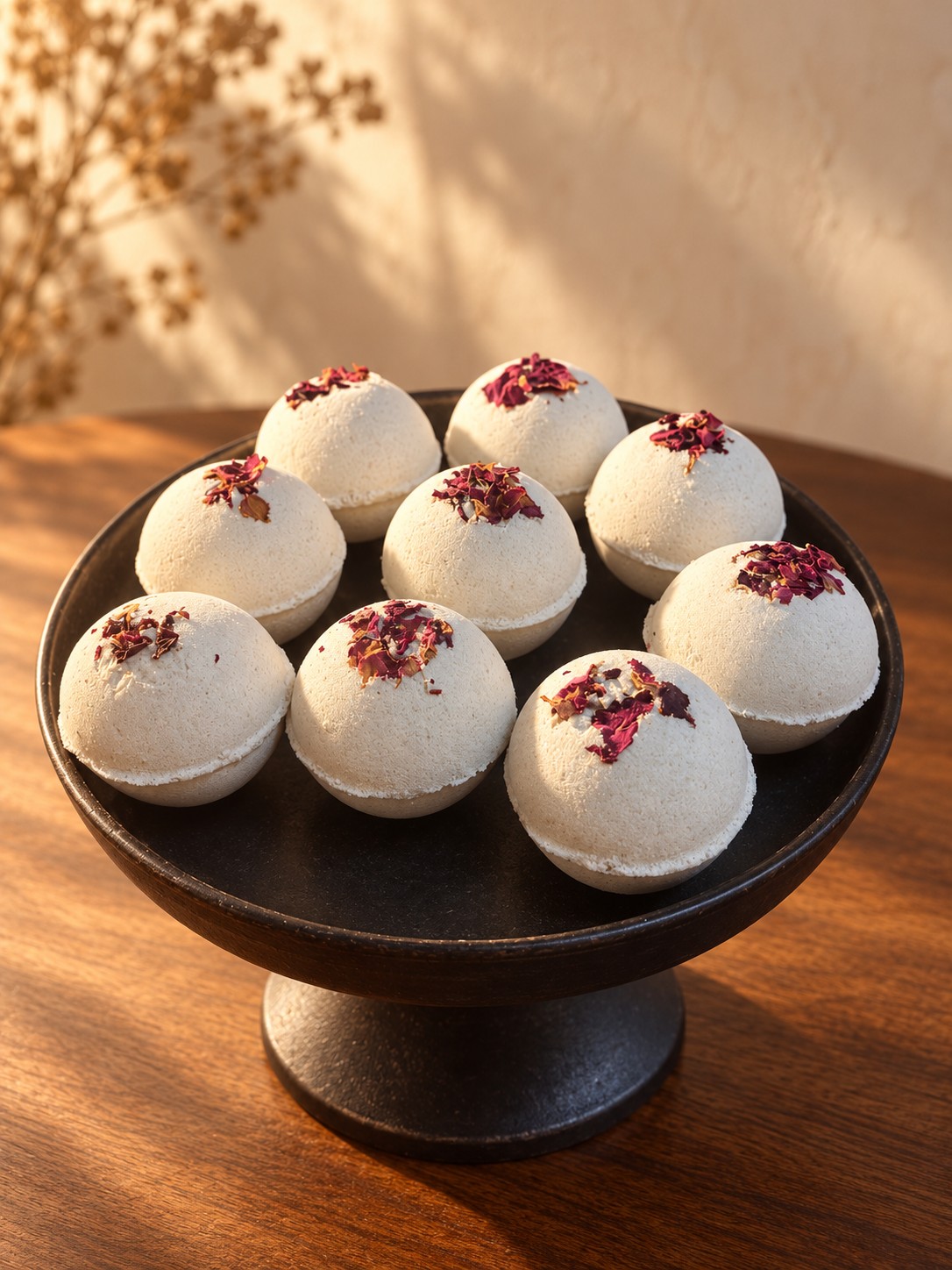 Nine ivory Moonlit Resin handmade bath bombs topped with dried rose petals on a black pedestal in warm natural light